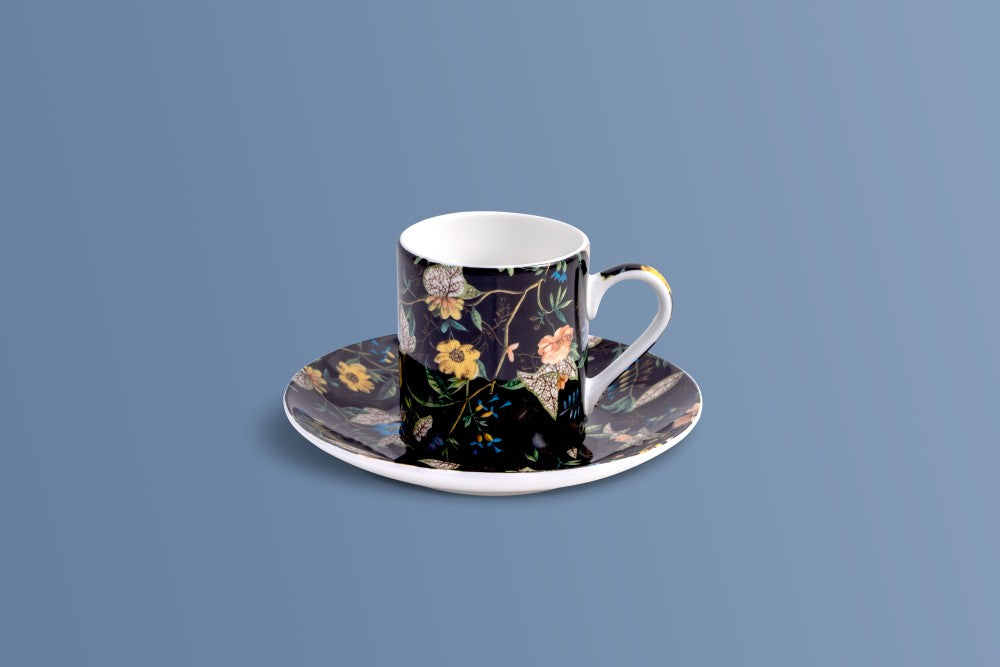 A ceramic coffee cup and saucer set with a black background and an intricate multicolor floral pattern, set against a solid blue-gray background.