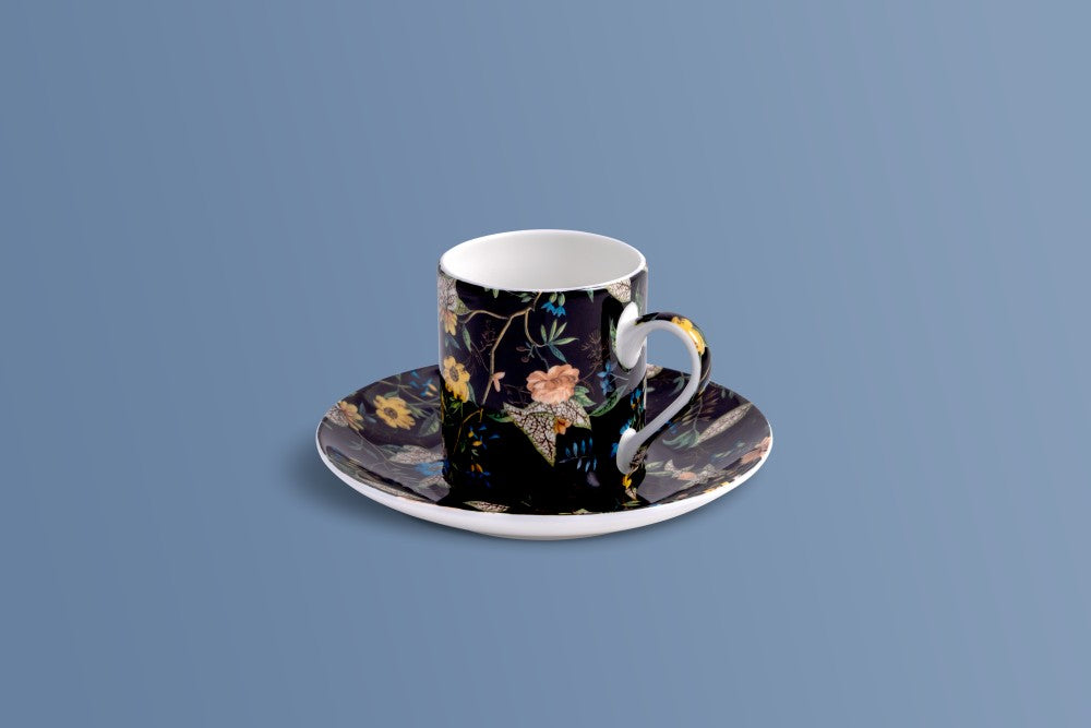 A single-piece coffee cup and saucer set with a black background and a detailed multicolor floral pattern, accented with gold leaf details on the handle and rim, set against a solid blue-gray background.