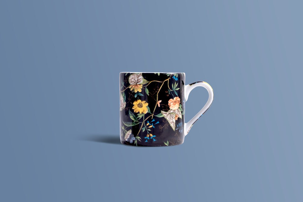 A white ceramic mug with a black background and a detailed floral pattern featuring yellow, orange, and blue flowers, set against a solid blue-gray background.