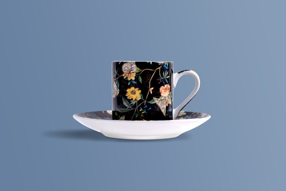 A white ceramic coffee cup with a black floral pattern and a matching white saucer, set against a solid blue-gray background.
