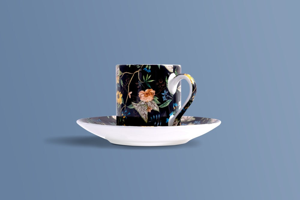 A ceramic coffee cup and saucer set on a solid blue-gray background. The cup features a dark navy floral pattern with orange, yellow, and green botanical details, while the saucer is solid white with a subtle dark rim.