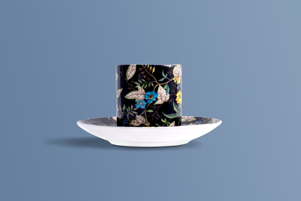 A tall cylindrical cup with a black background and an intricate botanical pattern of beige, blue, and yellow leaves and flowers, shown on a matching white saucer against a solid slate blue background.