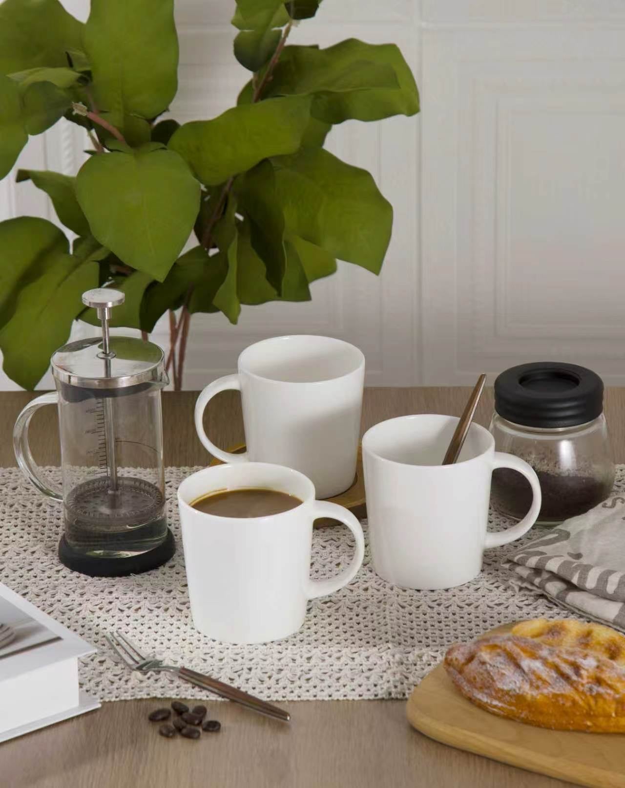White mugs with coffee, a French press, and a pastry on a table with a plant in the background.