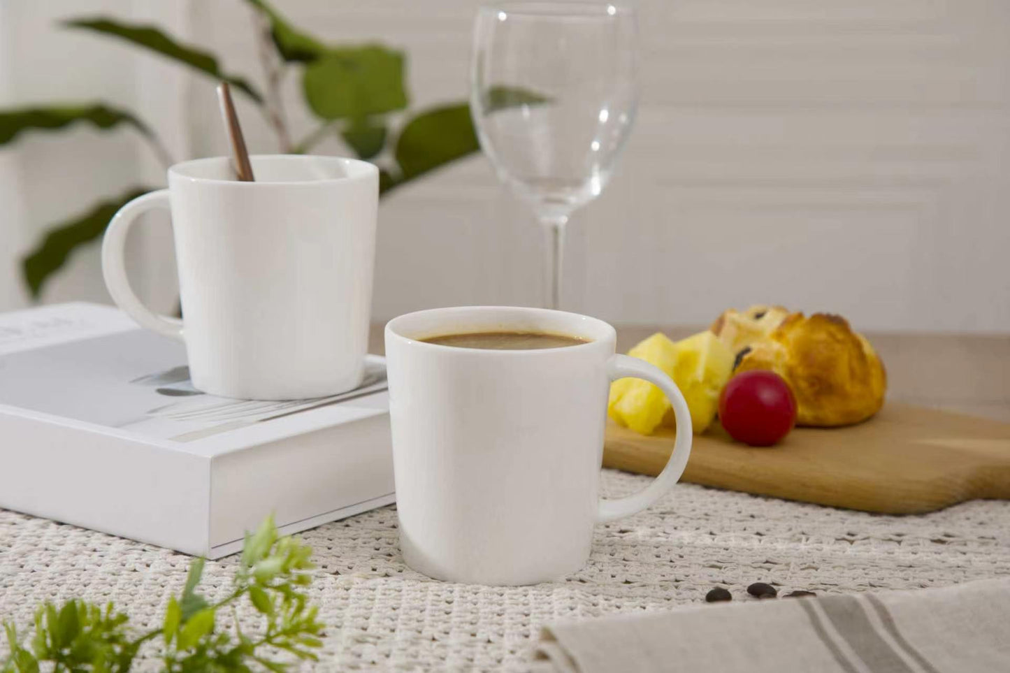 Two white mugs with coffee and a pastry on a table with a book and glass in the background.