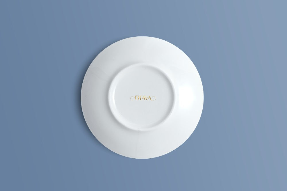 A white ceramic saucer shown from a top-down view, featuring the brand name Otava printed in gold in the centre, set against a solid blue-gray background.