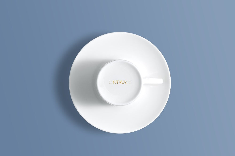 A white ceramic teacup and saucer set photographed from above on a muted blue background. The inside of the cup is printed with the brand name 'OTAVA' in gold.