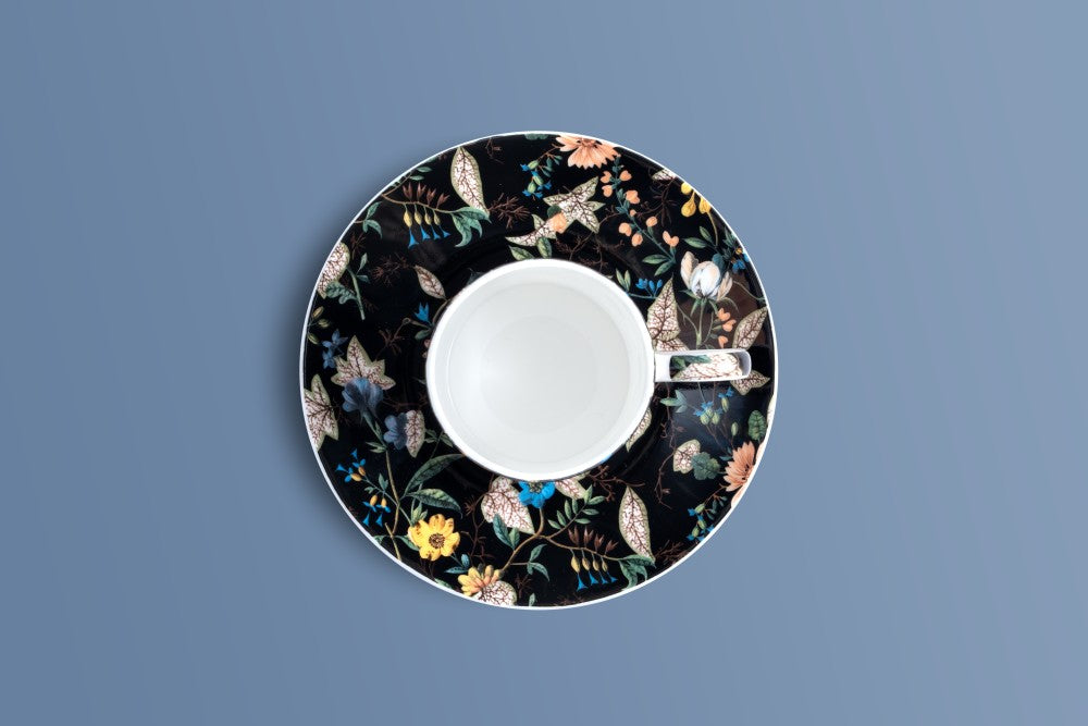 A white ceramic teacup and saucer set, viewed from directly above, featuring a black floral patterned saucer with a white cup in the center, set against a solid slate blue background.