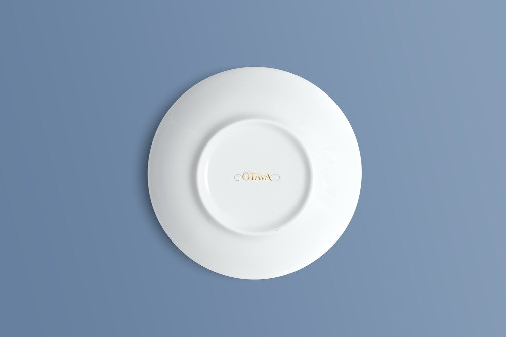 A white ceramic saucer shown from a top-down view, featuring a small gold logo in the centre, set against a solid blue background.