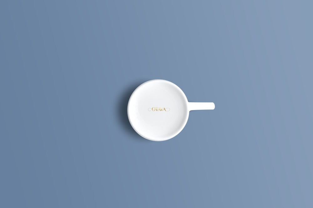 A white ceramic cup with a small handle, photographed from directly above on a solid blue background. The inside of the cup features the brand name 'Oria' printed in gold.