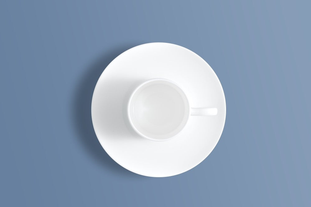 A minimalist white ceramic cup and saucer set, viewed from directly above on a solid slate blue background.