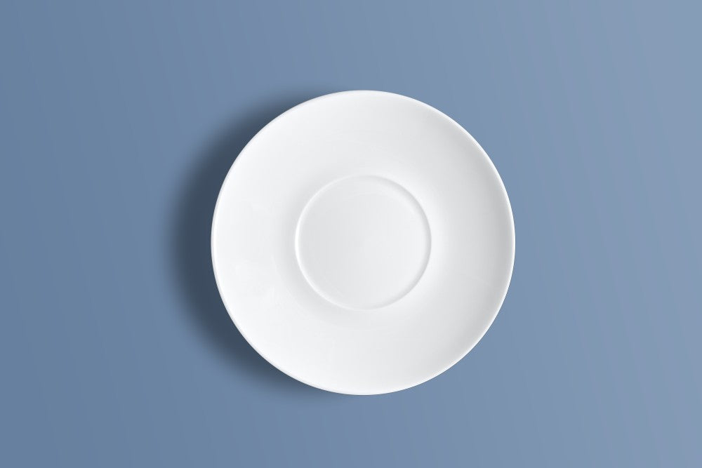 A white ceramic saucer with a raised centre ring, photographed from directly above on a solid slate blue background.