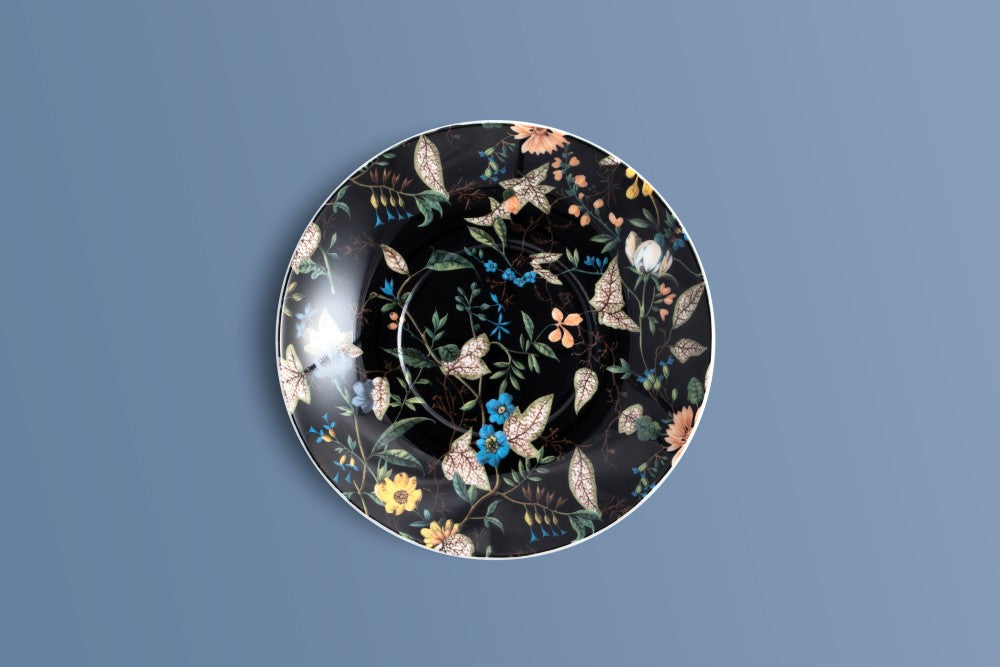 A round ceramic saucer with a black background and an intricate botanical pattern of leaves, flowers, and birds in muted earthy tones, blue, and yellow, shown against a solid slate blue background.