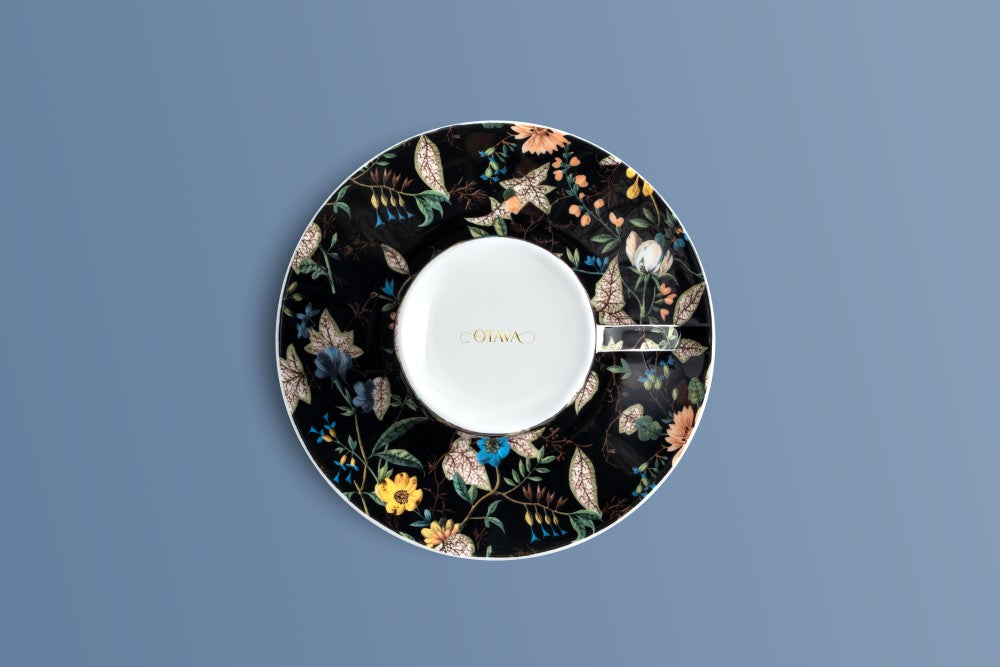 A circular ceramic saucer with a floral pattern on a black background, featuring a white centre and gold 'Otava' branding, set against a solid blue-gray background.
