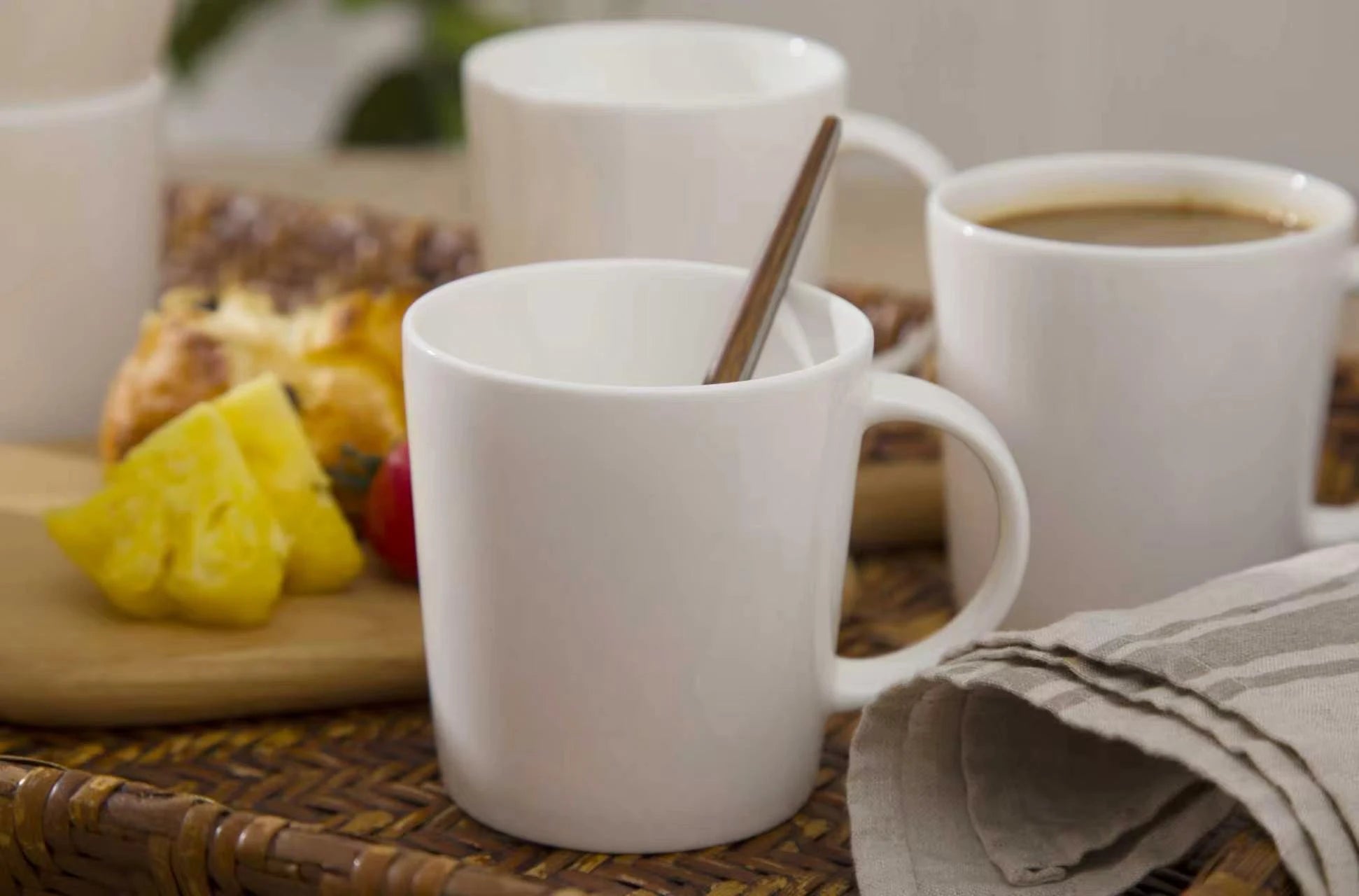 Two white mugs with a spoon on a table with a plate of food in the background